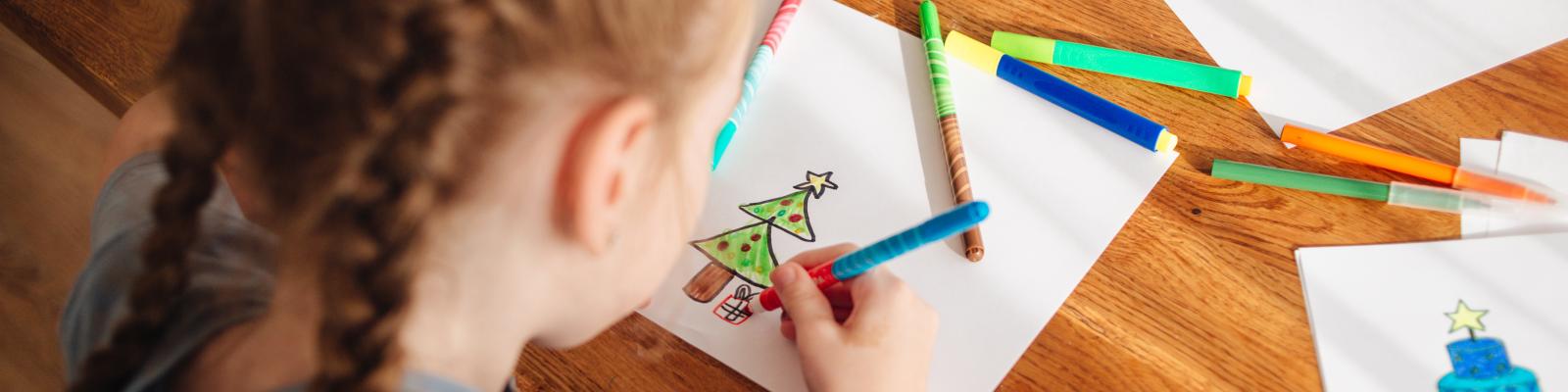 Child drawing a Christmas tree on a piece of paper with coloured pens around