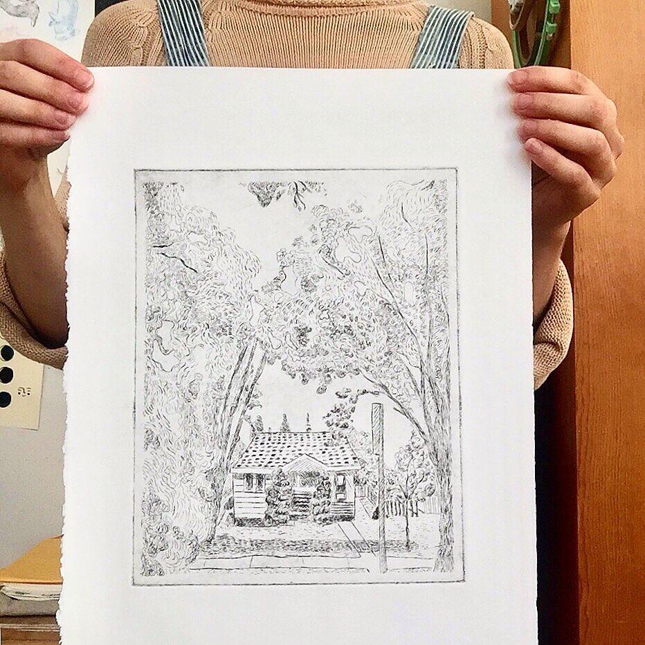 Person holding print of sketch. The sketch has a woodland cottage surrounded by trees.