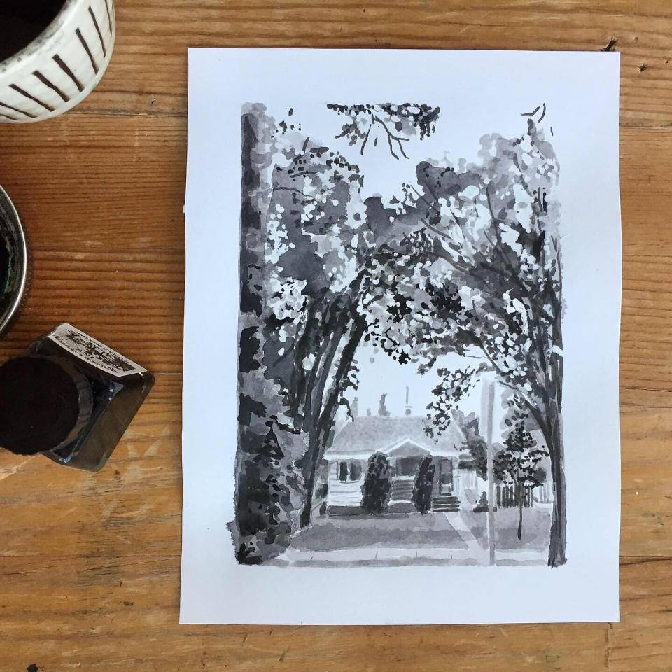 Ink drawing displayed on wooden desk. The ink drawing is of a cottage home surrounded by trees.