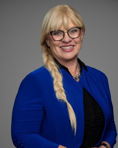 Headshot of Councillor Nancy McCurrach. There is a smiling blonde woman wearing glasses and a blue blouse. The background behind the councillor is grey.