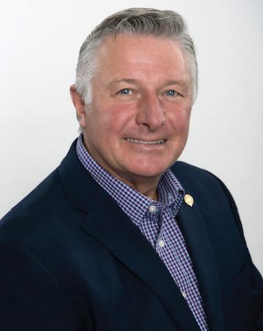 Headshot of Councillor Darrell Penner. There is a smiling man wearing a suit. The background behind the councillor is light grey.