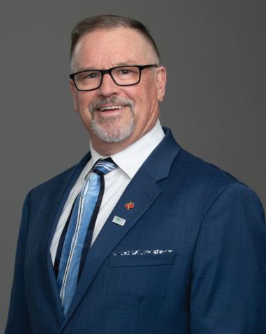 Headshot of Councillor Glenn Pollock. There is a smiling man wearing glasses and a blue suit and and tie. The background behind them is grey.