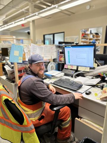 Headshot of Britton Ayers at their workstation. There is a man wearing a cap and dressed in overalls.