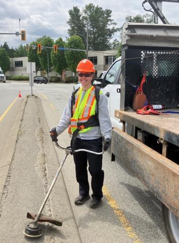 Public worker operating a weed whacker. They are wearing protective gear while standing near a truck.