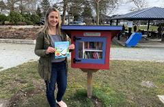 A woman holding a book standing in front of a little reading library in a park
