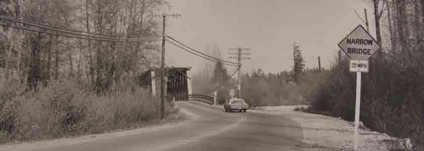 Car driving down a road towards a bridge. There is a sign warning about a narrow bridge ahead.